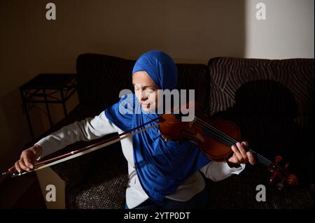 Eine heitere Szene einer Frau, die einen blauen Hijab trägt und geschickt Geige spielt. Das Setting ist intim mit einem Schwerpunkt auf ihrem Ausdruck und dem Instrumentarium Stockfoto