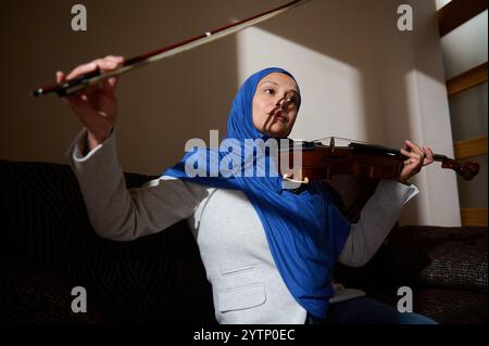 Eine Frau, die einen blauen Hijab und einen grauen Blazer trägt, spielt gekonnt Geige und konzentriert sich dabei auf ihre Leistung in einem warm beleuchteten Innenraum. Stockfoto