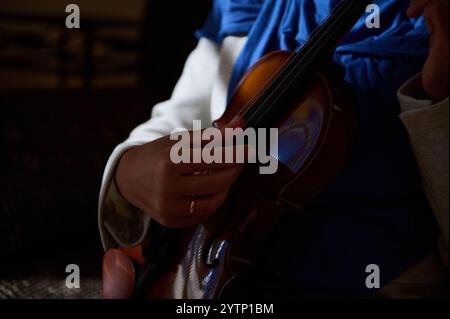 Eine detaillierte Ansicht der Hände, die anmutig eine Geige halten, vor einem sanft beleuchteten Hintergrund, was ein Gefühl musikalischer Leidenschaft und Kunstfertigkeit weckt. Stockfoto
