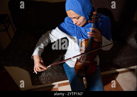 Eine junge Frau in einem blauen Hijab übt Geige auf einem Sofa und konzentriert sich darauf, ihre Musik in einer ruhigen Umgebung zu perfektionieren. Stockfoto