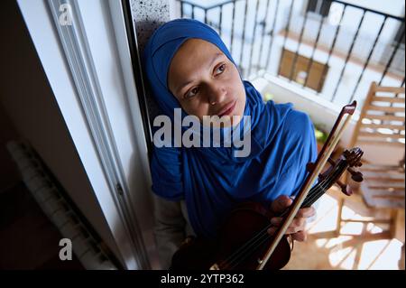 Eine Frau mit blauem Hijab steht auf einem Balkon, hält eine Geige und sieht besinnlich aus. Das Bild vermittelt Themen wie Musik, Reflexion und Kultu Stockfoto