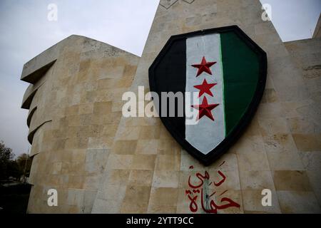 Hama, Syrien. Dezember 2024. Die syrische Oppositionsflagge ist auf einer adlerförmigen Mauer in Hama gemalt, nachdem Oppositionskämpfer die Stadt von den syrischen Regierungstruppen eingenommen haben. Seit dem 27. November sind syrische Oppositionsfraktionen unter der Führung von Hayat Tahrir al-Sham rasch aus ihrer Festung in Idlib vorgezogen und haben große strategische Gebiete von der syrischen Regierung von Baschar Al-Assad eingenommen. Die Rebellen haben ihre laufende Militäroperation als Abschreckung der Aggression bezeichnet: IMAGESLIVE/Alamy Live News Stockfoto