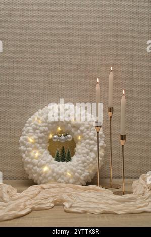 Weihnachtliche Einrichtung, weicher Plüschkranz mit Lichtern, drei Kerzen in goldenem Halter, beigefarbenes Tuch auf Holztisch Stockfoto