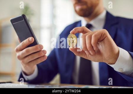 Börsenhändler oder Geschäftsmann, der am Schreibtisch sitzt und Handy und Bitcoin in der Hand hält. Stockfoto