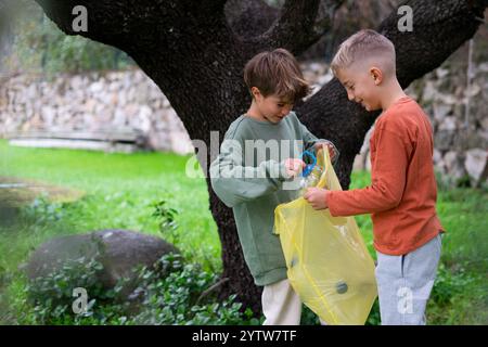 Zwei Kinder legen Plastikflaschen in einen gelben Müllbeutel in einem Park, um Umweltbewusstsein und Recycling zu fördern Stockfoto