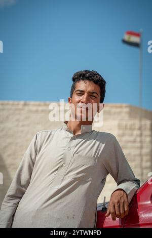 Porträt eines ägyptischen Taxifahrers, der neben seinem Fahrzeug steht, mit der ägyptischen Flagge im Hintergrund auf dem deutschen Kriegsfriedhof El Alamein. Stockfoto