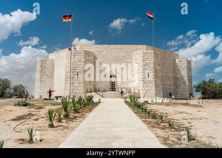 Der deutsche Kriegsfriedhof El Alamein steht wie eine Festung unter blauem Himmel, mit Flaggen von Deutschland und Ägypten oben und Grün säumen den Wüstenweg Stockfoto