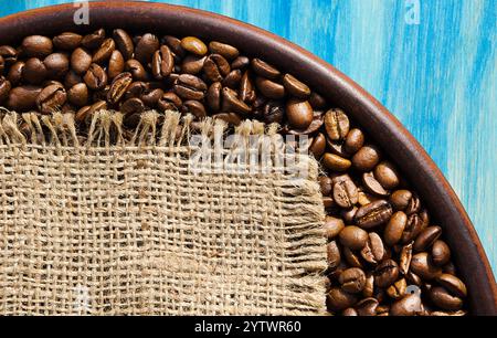 Geröstete Kaffeebohnen auf einer Keramikplatte auf türkisfarbenem Hintergrund. Ein Stück Sackleinen für Text. Körner einer tropischen Pflanze. Stockfoto