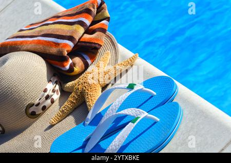 Flipflops, ein großer Seestern, ein panamahut und ein farbenfrohes Handtuch neben dem Pool. Sommerferienkonzept vor dem Hintergrund des blauen Wassers. Stockfoto