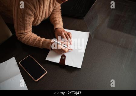 Eine Person, die einen Pullover trägt, schreibt auf einem Papier neben einem Smartphone auf einem Schreibtisch. Dieses Bild erfasst einen fokussierten Moment der Notizen oder Planung in einem Comfo Stockfoto