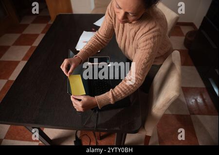 Eine Frau in einem kuscheligen Pullover legt eine gelbe Haftnotiz auf ihren Laptop und fokussiert sich intensiv. Stockfoto