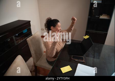 Eine freudige Frau hebt die Arme zur Feier und reagiert begeistert auf gute Nachrichten auf ihrem Laptop, während sie von zu Hause aus arbeitet. Stockfoto