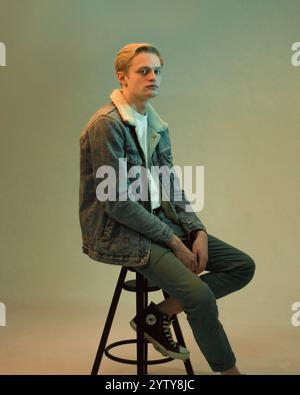 Der junge blonde Mann in einer Jeansjacke sitzt in voller Wucherung auf einem Stuhl im Studio Stockfoto