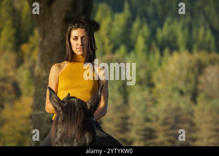 Eine entschlossene Frau in senfarmem ärmellosem Oberteil sitzt auf einem schwarzen Pferd mit einer ruhigen Landschaft mit üppigen grünen Bäumen und sanftem natürlichem Licht. Stockfoto