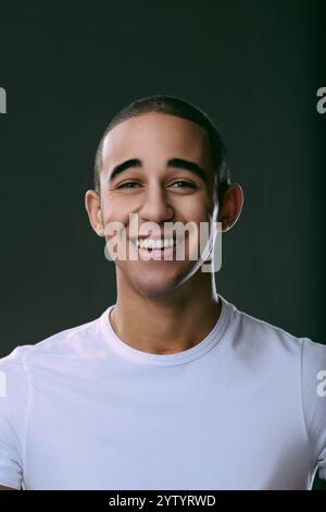 Fröhlicher junger Mann, der breit lächelt, seine Zähne vor dunklem Hintergrund zeigt und Glück und Selbstvertrauen in einem Studio ausstrahlt. Hochformat Stockfoto