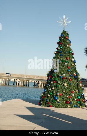 Vertikaler Blick auf einen rot, weiß und grün dekorierten Weihnachtsbaum mit Kugeln. Neben Boca Ciega Bay hinten. In St Pete Beach, FL. Blauer Himmel und ruhiges Wasser. Stockfoto