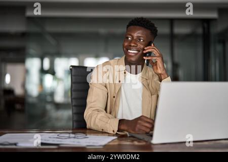 Glücklicher afrikanischer Geschäftsmann, der auf dem Handy spricht und im Büro am Laptop arbeitet. Stockfoto