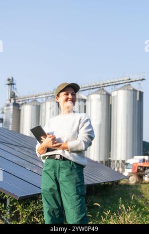 Farmerin mit digitalem Tablet auf einem modernen Bauernhof mit Solarpaneelen. Landwirtschaftliche Silos im Hintergrund. Stockfoto