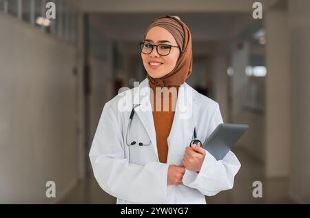 Porträt der glücklichen muslimischen Ärztin oder Krankenschwester mit weißem Mantel mit Stethoskop im Krankenhausflur mit digitalem Tablet, das in die Kamera schaut und Stockfoto