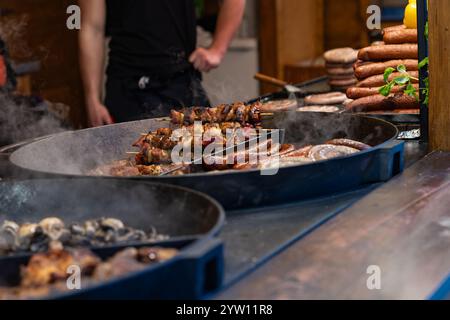 Köstliches Street Food auf dem Weihnachtsmarkt mit gegrillten Würstchen, Fleischspießen und Pilzen, die auf großen gusseisernen Pfannen brutzeln Stockfoto