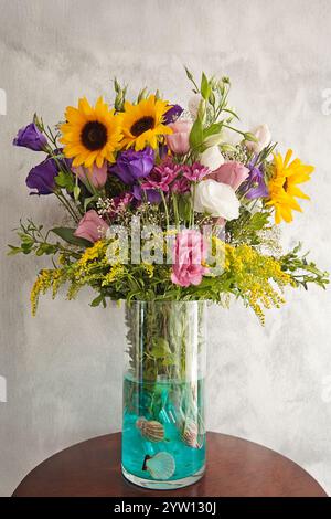 Ein leuchtender Blumenstrauß aus Sonnenblumen, lila Lisianthus, rosa Rosen und gelben solidago, in einer Glasvase gefüllt mit blauem Wasser und Muscheln, Stockfoto