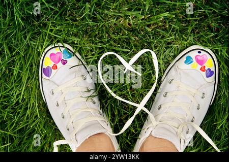 Ein Paar weiße Sneakers mit farbenfrohen Herzmotiven, die auf leuchtendem grünem Gras ruhen und Schnürsenkel eine Herzform bilden. Stockfoto