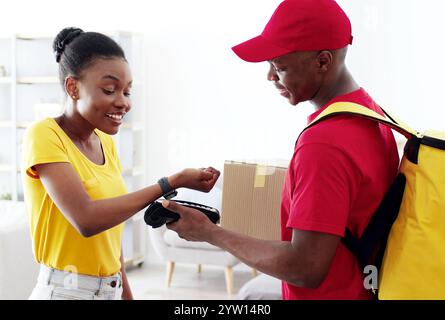 Intelligente drahtlose Bezahlung. afroamerikanerin, die mit Smartwatch für die Bestellung bezahlt, Kurier mit Pos-Terminal Giving Box Stockfoto