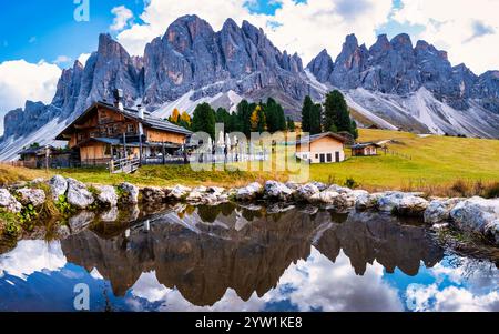 Atemberaubende Gipfel der Dolomiten erheben sich majestätisch gegen einen blauen Himmel und spiegeln sich wunderschön in einem ruhigen Teich. Holzhütten im üppigen greene Stockfoto