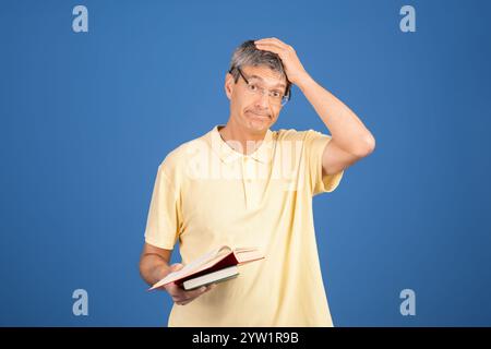 Reifer Mann, der verwirrt aussieht, während er ein Buch vor blauem Hintergrund hält Stockfoto