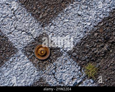 Zwischen sich kreuzenden weißen Linien befindet sich ein Messmarker aus Metall in Asphalt Stockfoto