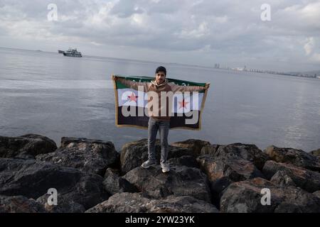 Izmir, Türkei. Dezember 2024. Ein syrischer Flüchtling posiert am Strand mit einer Flagge der syrischen Nationalarmee. Der Sturz der Regierung von Baschar al-Assad mit dem Einzug von Hayat Tahrir al-Sham nach Damaskus hat Millionen syrischer Flüchtlinge, die in der Türkei leben, auf die Straße gebracht. Die in Izmir lebenden Syrer gingen auf die Straße und zeigten ihre Freude. Quelle: SOPA Images Limited/Alamy Live News Stockfoto