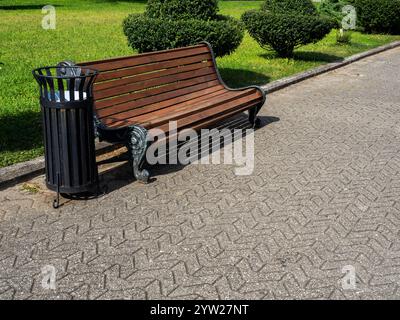 Leere Vintage Holzbank mit schmiedeeisernen Beinen in der Nähe des Mülltonnen auf dem grauen Weg in der Nähe von grünem Gras und Bäumen im Park an sonnigen Tagen. Stockfoto