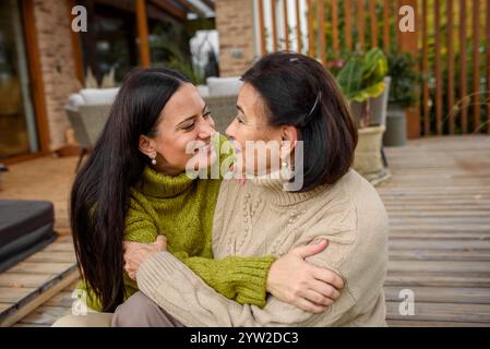 Ältere Tochter mit älterer Mutter, die auf einer hölzernen Terrasse sitzt und sich umarmt. Muttertagskonzept. Stockfoto