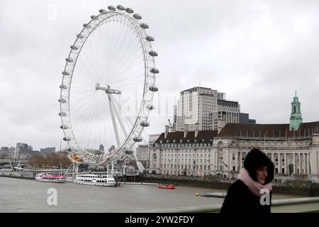 London, Großbritannien. Dezember 2024. Eine Frau geht am 8. Dezember 2024 auf der Westminster Bridge mit dem London Eye im Hintergrund. Zehntausende von Häusern wurden ohne Strom gelassen, Bahn- und Luftverkehr wurden gestört und Sportveranstaltungen in ganz Großbritannien wurden am Samstag abgesagt, als der Sturm Darragh zuschlug. Quelle: Li Ying/Xinhua/Alamy Live News Stockfoto