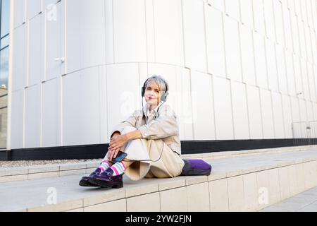 Lächelnde, Trendige Reife Frau, Die Musik Über Kopfhörer Hört, Während Sie Auf Den Stufen Des White Modern Building In Der Stadt Sitzt. Stockfoto