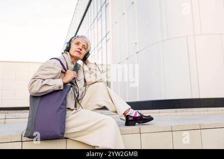 Schöne trendige Reife Frau mit kurzen Haaren und Kopfhörern in Beige Kleidung sitzt draußen in der Stadt, hört Musik, Podcast oder Hörbuch Agai Stockfoto