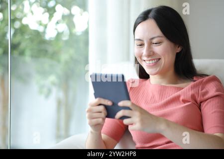 Glückliche asiatische Frau, die eBook liest und zu Hause auf einer Couch sitzt Stockfoto