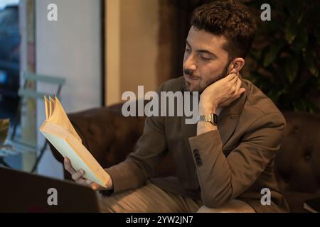 Stilvoller junger Mann in braunem Blazer, der Buch im stimmungsvollen Café liest. Professionelle Genießen Sie friedliche Momente der Literatur mit warmer Beleuchtung und Stockfoto