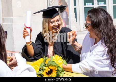 Ein lächelnder Absolvent in schwarzer Mütze und Kleid mit Diplom und Sonnenblumen feiert mit einer Frau im Freien, Bournemouth, Dorset UK Stockfoto