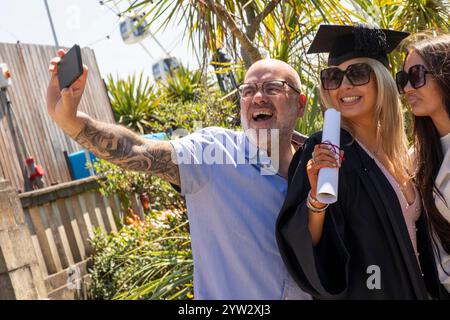 Eine fröhliche Familie, die den Abschlussfeiertag mit einem Selfie feiert, mit einer jungen Frau in Mütze und Kleid, die ihr Diplom hält, Bournemouth, Dorset UK Stockfoto