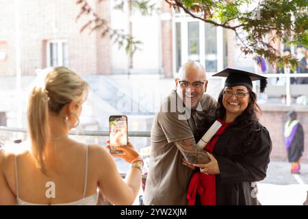 Lächelnde Frau in Abschlussmütze und Kleid posiert für ein Foto mit einem Mann, während eine andere Person ihr Foto mit einem Smartphone macht, Bournemouth, Dorset UK Stockfoto