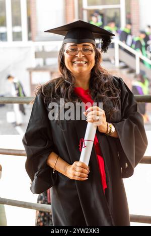 Lächelnde Frau in Abschlusskleidung, die ein Diplom hält und stolz mit Absolventen im Hintergrund steht, Bournemouth, Dorset, Großbritannien Stockfoto