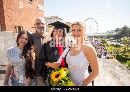 Familie feiert eine Abschlusszeremonie mit einer lächelnden Frau in Mütze und Kleid, die ein Diplom hält, mit einem Riesenrad im Hintergrund, Bournemouth, Dorset UK Stockfoto