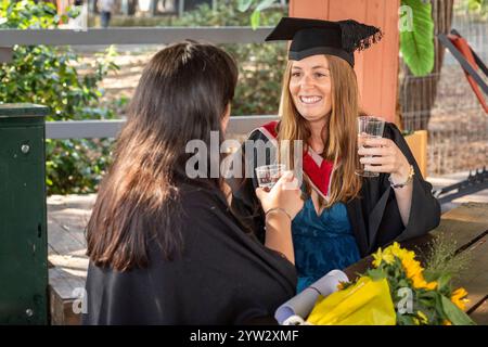 Lächelnder Absolvent in Mütze und Kleid, der mit einem Freund bei einem Drink in einem Outdoor-Veranstaltungsort in Bournemouth, Dorset, Großbritannien, feiert Stockfoto