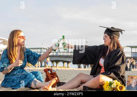 Zwei Frauen feiern am Strand mit einem Toast, eine in Abschlusskleidung und ein Blumenstrauß in der Nähe, Bournemouth, Dorset UK Stockfoto