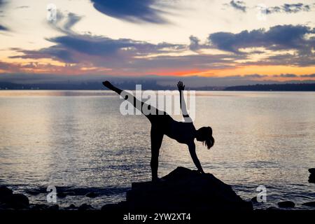 Silhouette einer Frau, die bei Sonnenuntergang auf einem Felsen am Ufer eine Yoga-Pose spielt. Stockfoto