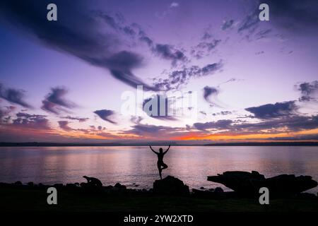 Silhouette einer Frau, die bei Sonnenuntergang auf einem Felsen am Ufer eine Yoga-Pose spielt. Stockfoto