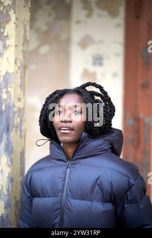 Eine selbstbewusste Frau mit schwarzen geflochtenen Haaren in einer geschwollenen schwarzen Jacke steht vor einer verblassten Wand in Brandenburg Stockfoto