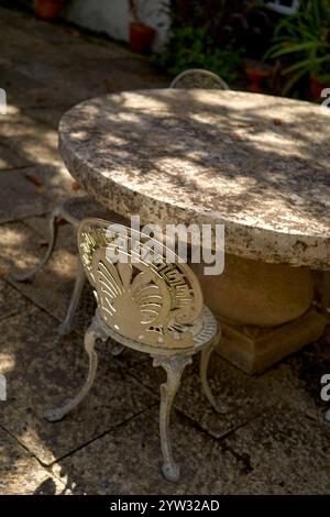 Sonnenlicht zieht durch die Blätter auf einen alten, verzierten Metallstuhl und einen runden Steintisch in einer ruhigen Gartenumgebung, Portugal Stockfoto