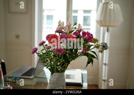 Frische bunte Blumen in einer weißen Vase auf einem Tisch mit Laptop und Lampe in einem hellen Raum. Stockfoto